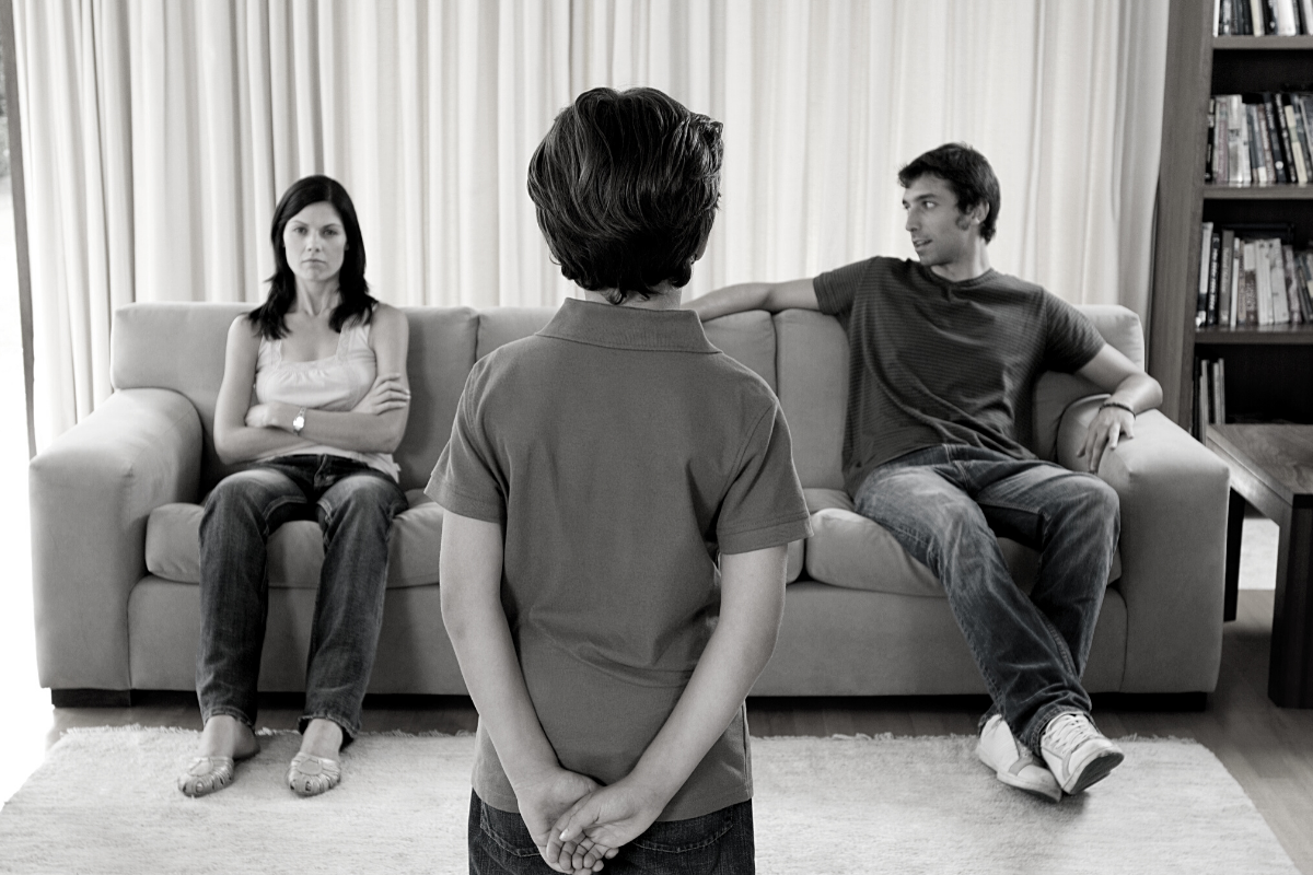 child standing with back to camera in front of angry mum and dad on sofa