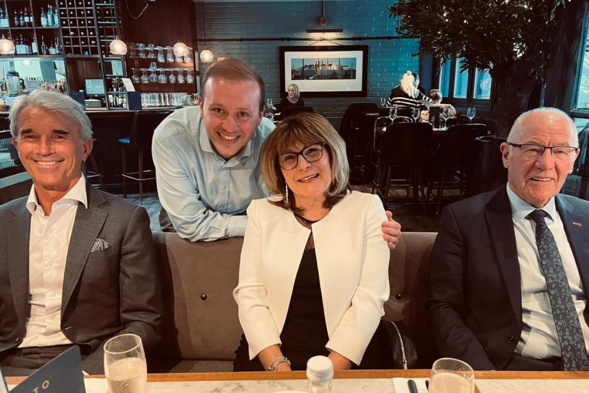 Andrea is sitting at a restaurant table with Ron Walker on the left and Keith Thompson to her right, Alistair Cartwright is leaning over behind her